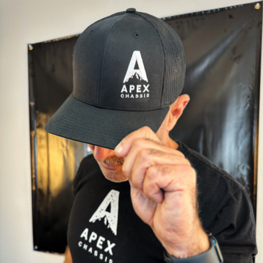 Wearing an Apex Chassis Apex SnapBack Trucker Hat with breathable mesh panels and a matching shirt, a person tilts the brim to partially hide their face against a dark background.